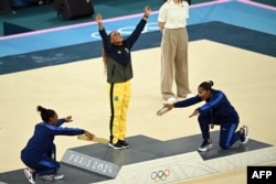 Симон Байлс (вляво) и Джордан Чайлс (вдясно) коленичат пред златната медалистка на земя в Париж 2024 Ребека Андраде, 5 август 2024 г.